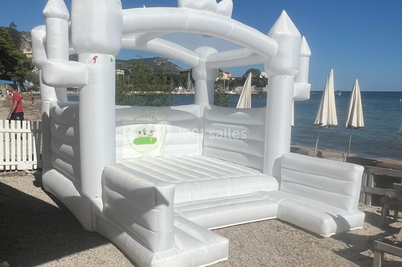 Château gonflable blanc installé en extérieur près d'une plage, avec parasols et mer en arrière-plan.
