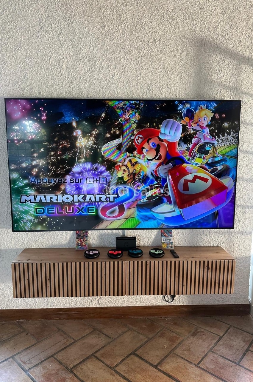 Écran affichant le jeu Mario Kart Deluxe avec des manettes posées sur un meuble en dessous.