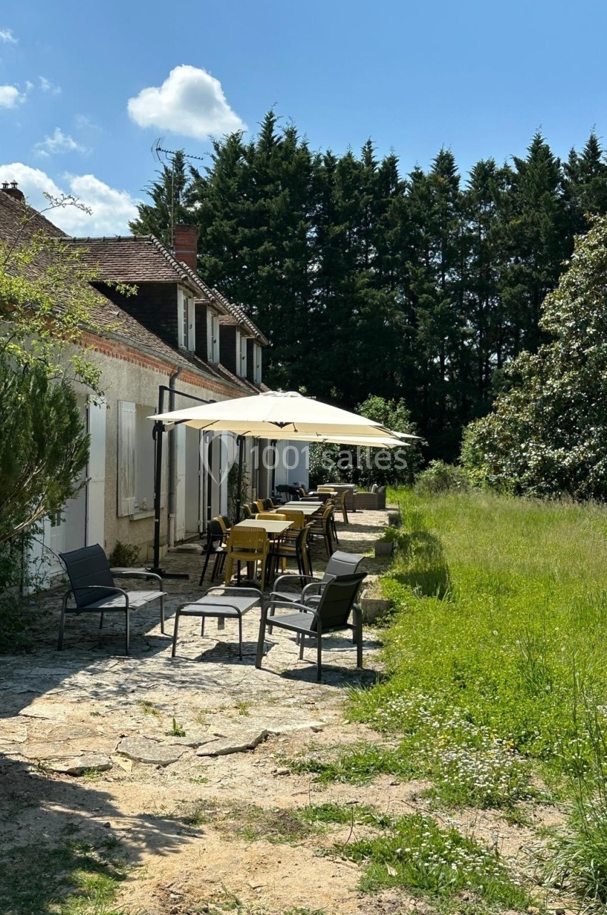 Terrasse ensoleillée avec tables, chaises et parasols devant une maison en pierre entourée de verdure.