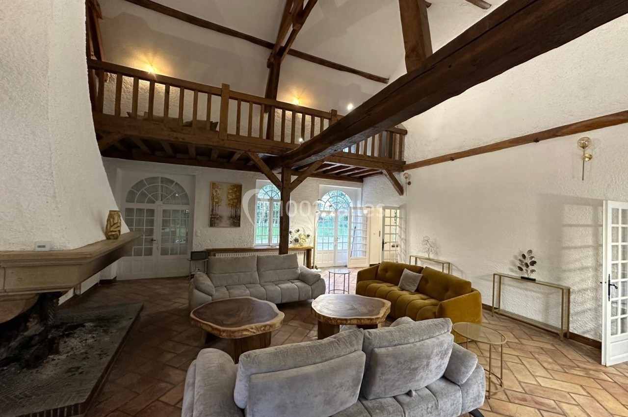 Salon spacieux avec poutres apparentes, cheminée, canapés gris et jaunes, tables en bois, et mezzanine en bois.