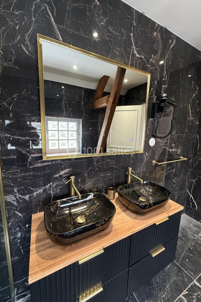 Salle de bain moderne avec mur en marbre noir, double vasque noire, robinetterie dorée et grand miroir rectangulaire.