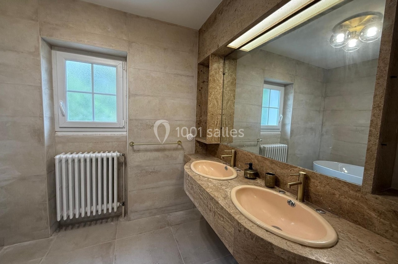 Salle de bain avec deux lavabos roses, un grand miroir, un radiateur blanc et une fenêtre à gauche.