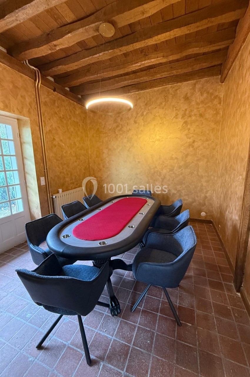 Table de poker rouge entourée de chaises noires dans une pièce aux murs texturés et au plafond en bois.