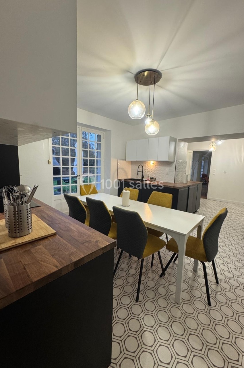 Cuisine moderne avec table à manger blanche, chaises jaunes et grises, plan de travail en bois et éclairage suspendu.