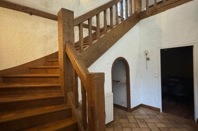 Escalier en bois avec rambarde, dans un intérieur aux murs blancs texturés et sol en carrelage.