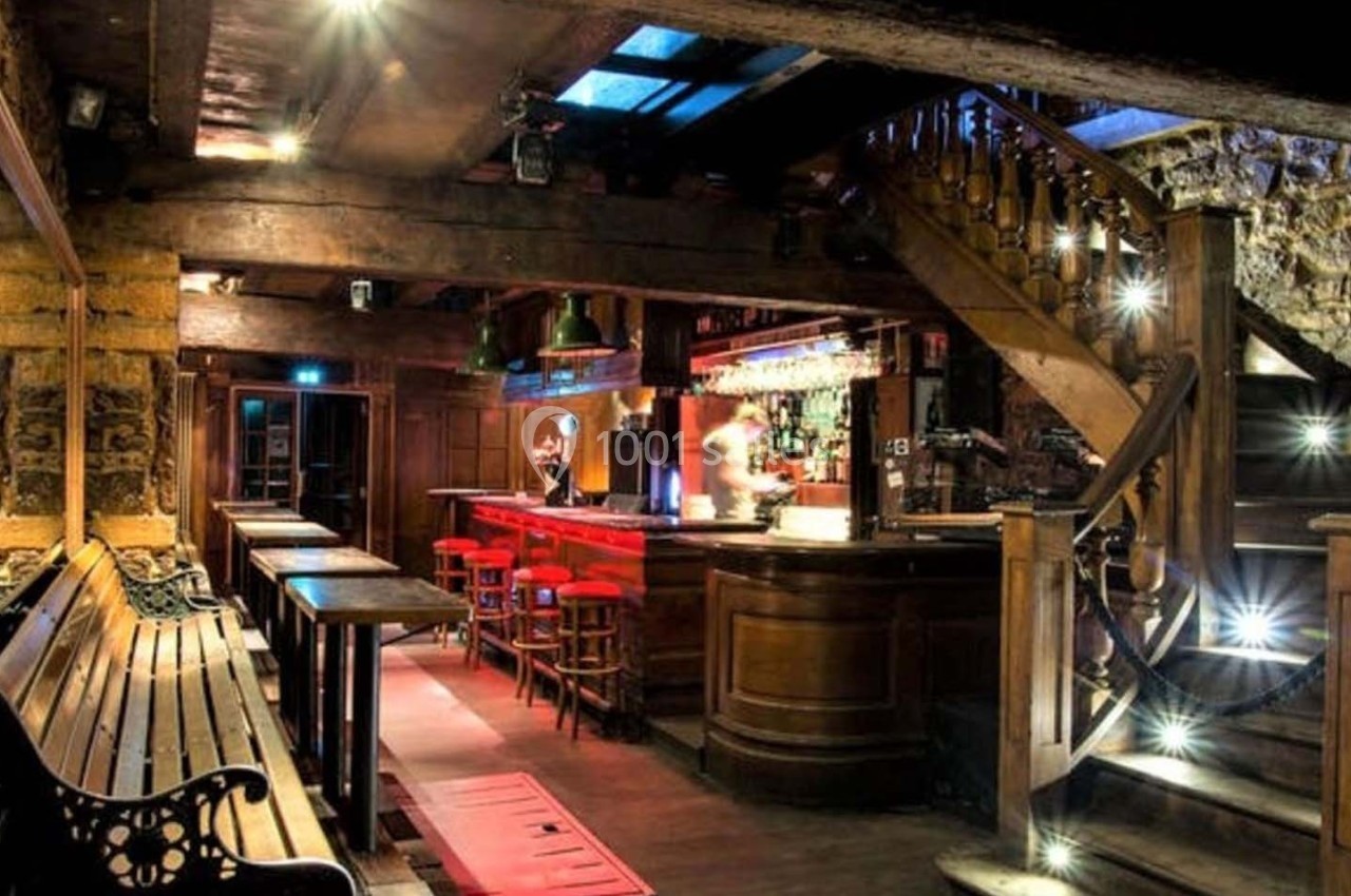 Intérieur d'un bar avec un comptoir éclairé, des tabourets rouges, un escalier en bois et des murs en pierre apparente.
