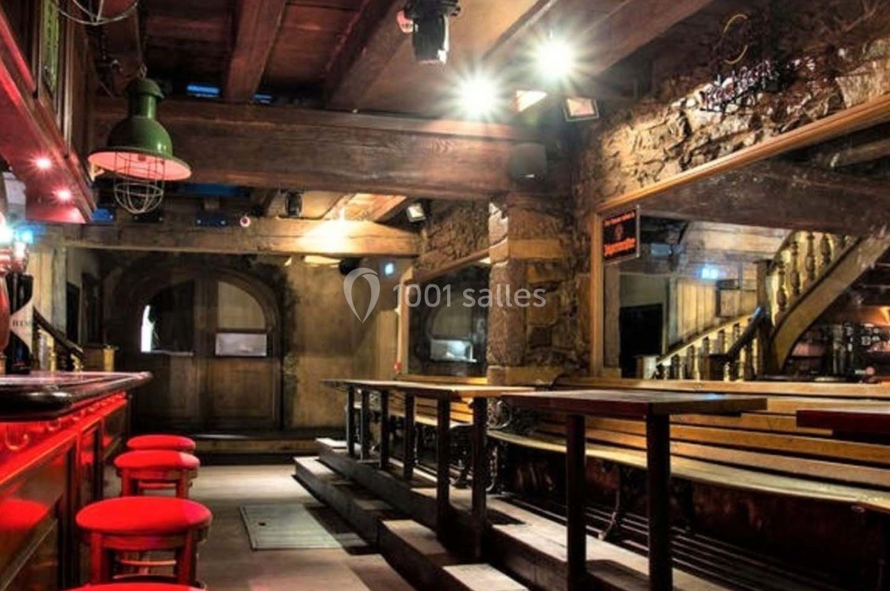 Intérieur d'un bar rustique avec des tabourets rouges, des poutres en bois et un éclairage tamisé.