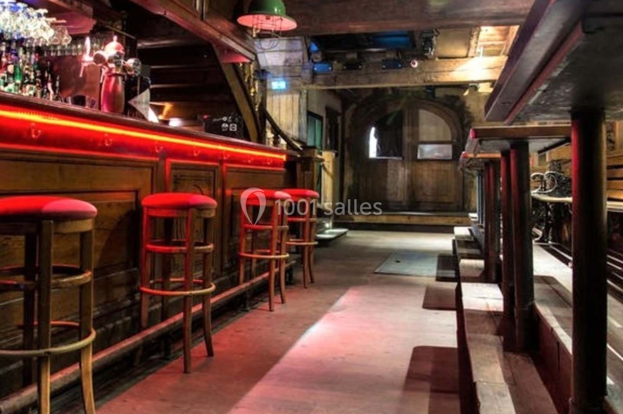Intérieur d'un bar avec comptoir en bois, tabourets rouges et éclairage tamisé.