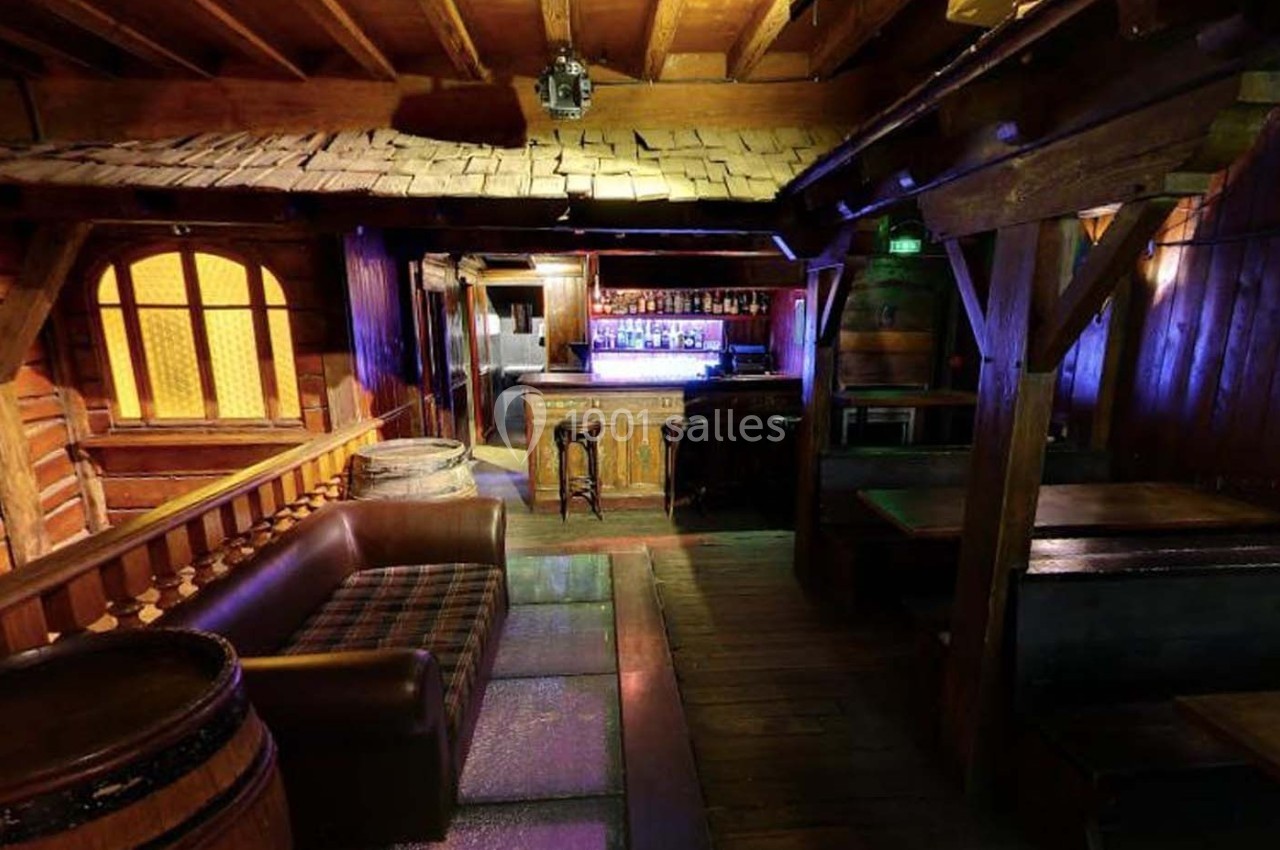 Intérieur d'un bar rustique avec mobilier en bois, canapé, tonneaux et comptoir éclairé par des lumières colorées.