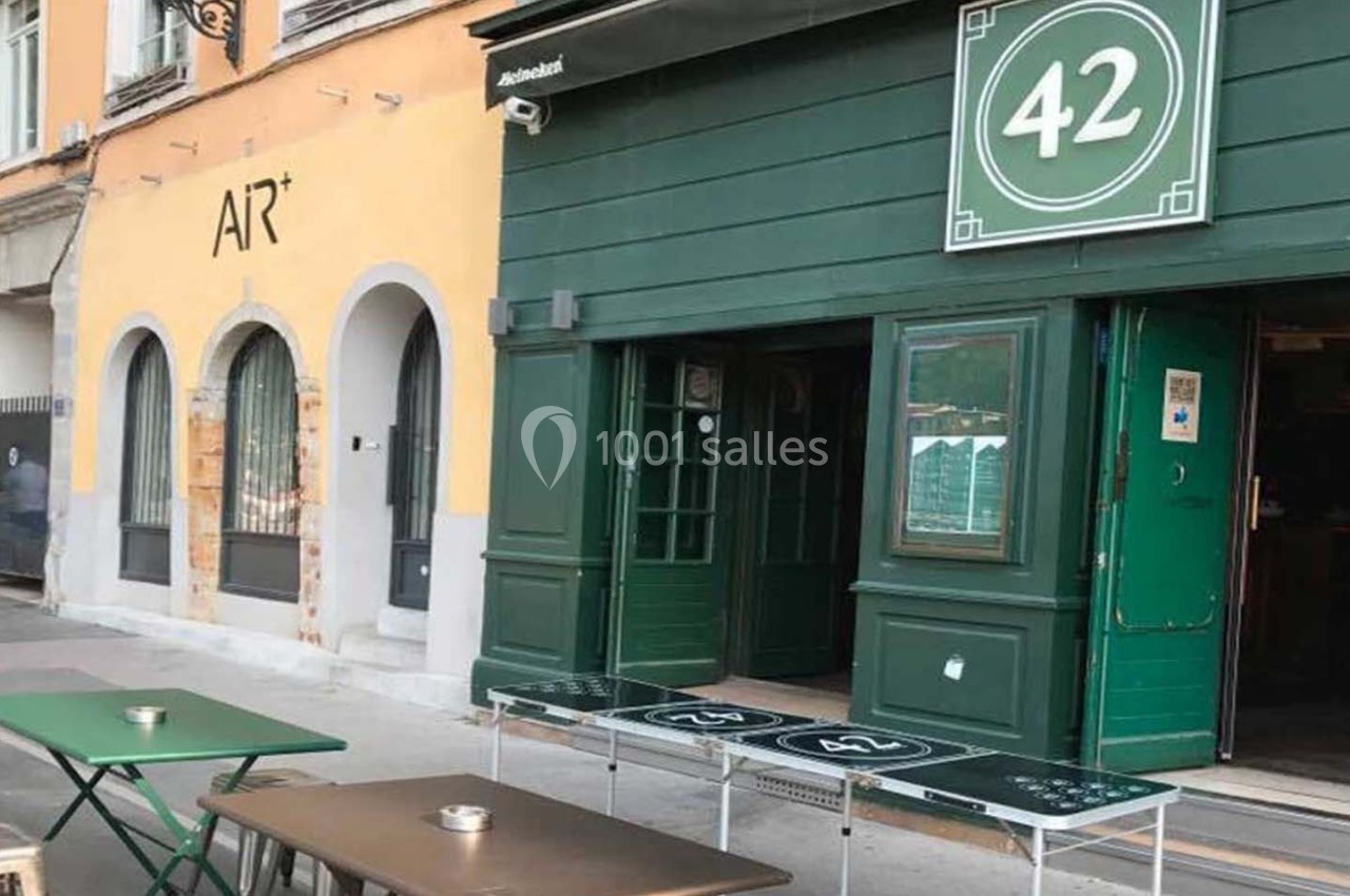 Façade d'un café avec terrasse, tables et bancs verts, situé entre des bâtiments colorés en rue piétonne.