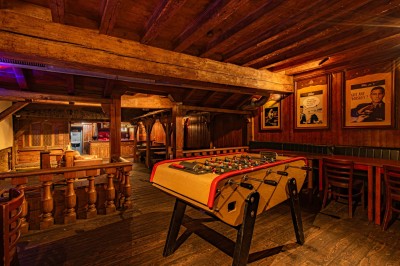 Miniature Salle du Haut Salle en bois chaleureux avec un baby-foot au centre, entourée de tables et chaises, éclairage tamisé.