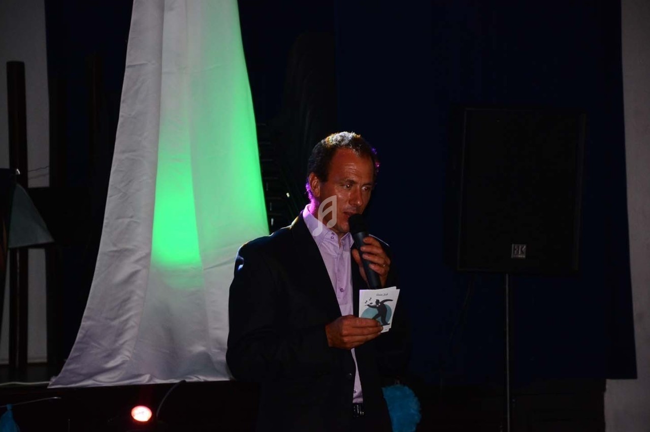 Un homme en costume parle dans un microphone, tenant une carte, devant un fond sombre avec un éclairage coloré.