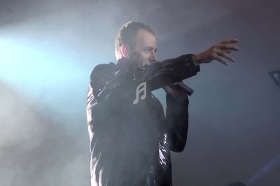 Un homme chante sur scène avec un micro, éclairé par une lumière rouge dans une ambiance sombre.