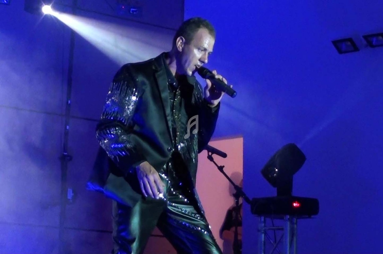 Un chanteur en costume noir brillant se produit sur scène sous un éclairage bleu avec un micro à la main.