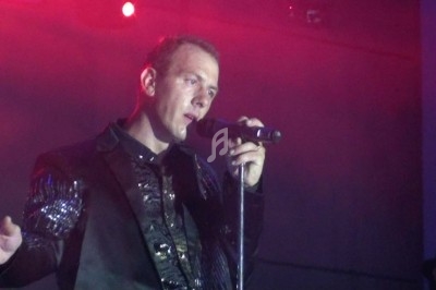 Un homme chante sur scène avec un micro, éclairé par une lumière rouge dans une ambiance de concert.