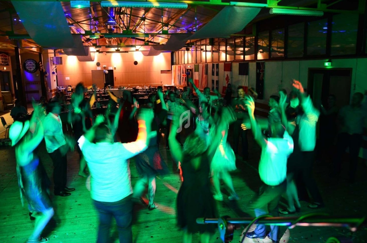 Des personnes dansent sous des lumières colorées dans une salle avec un plafond en bois et des tables en arrière-plan.