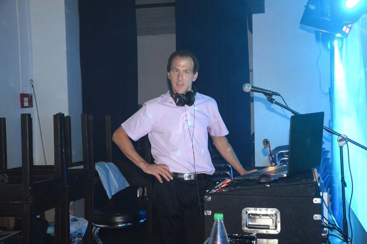 Un homme debout derrière une table de mixage avec un ordinateur et un micro, dans une salle éclairée en bleu.