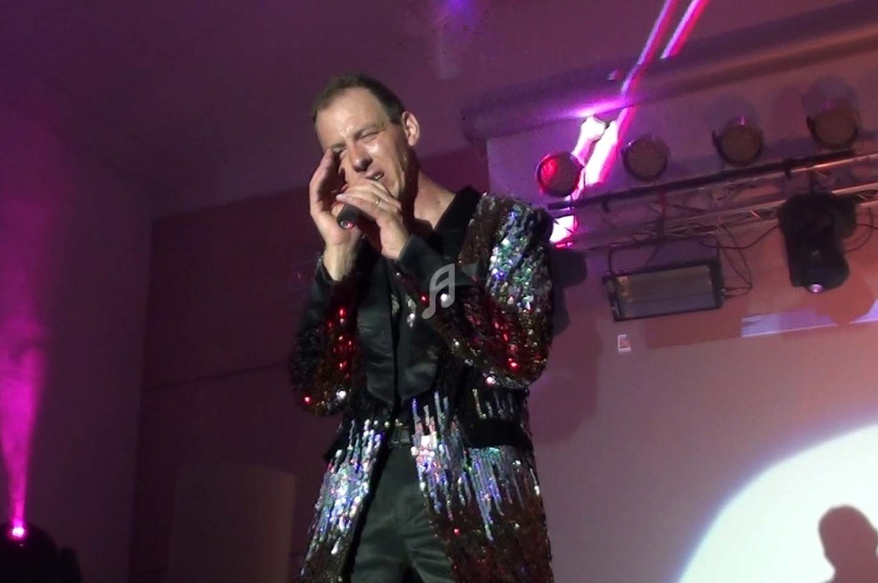 Un chanteur en veste à paillettes interprète sur scène, éclairé par des projecteurs colorés.