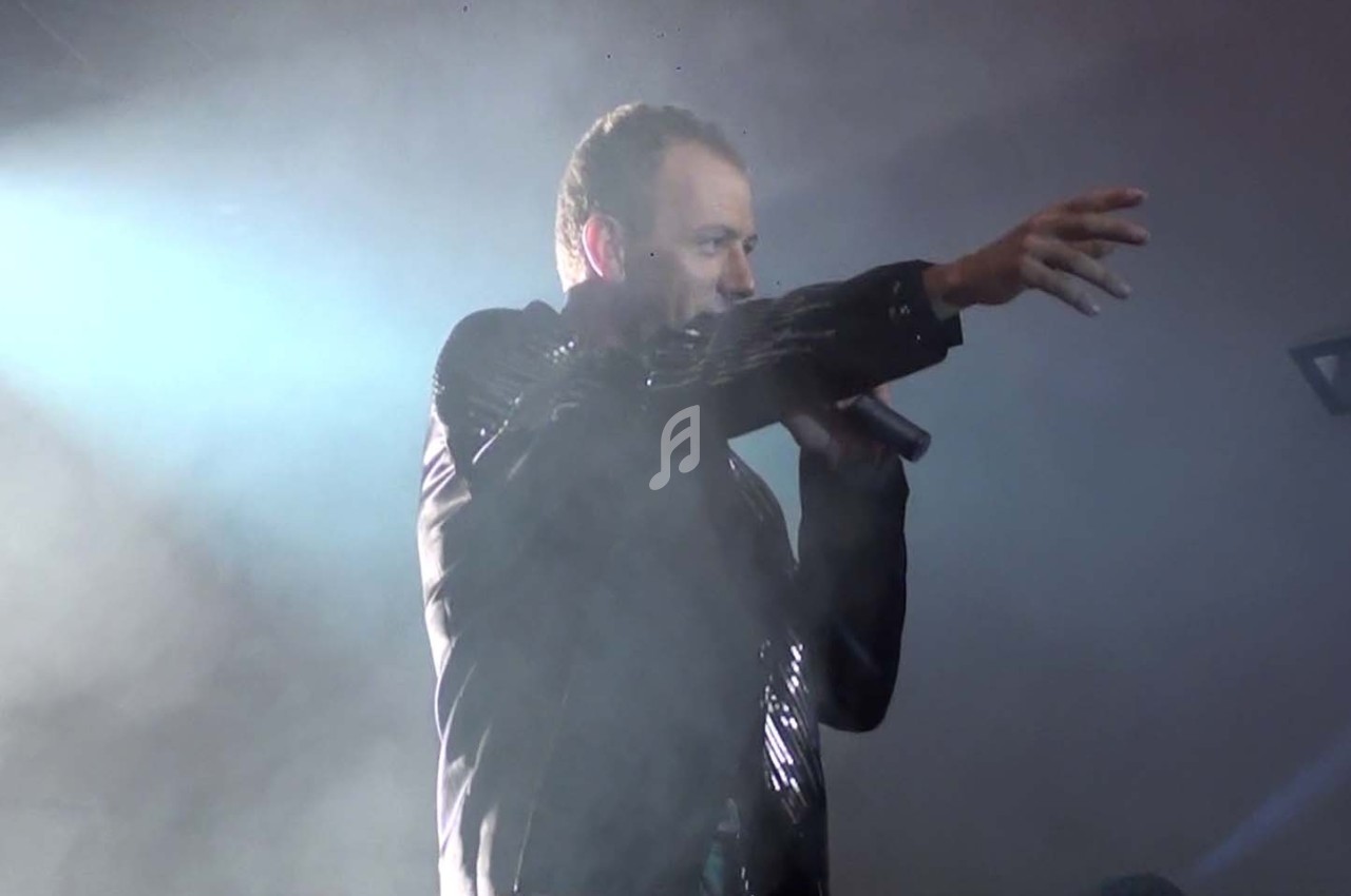 Un chanteur en veste noire tient un micro et pointe du doigt sur une scène éclairée par des projecteurs.