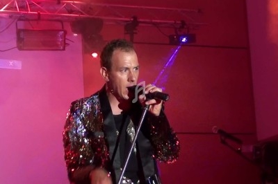 Un homme chante sur scène avec un micro, éclairé par une lumière rouge dans une ambiance de concert.