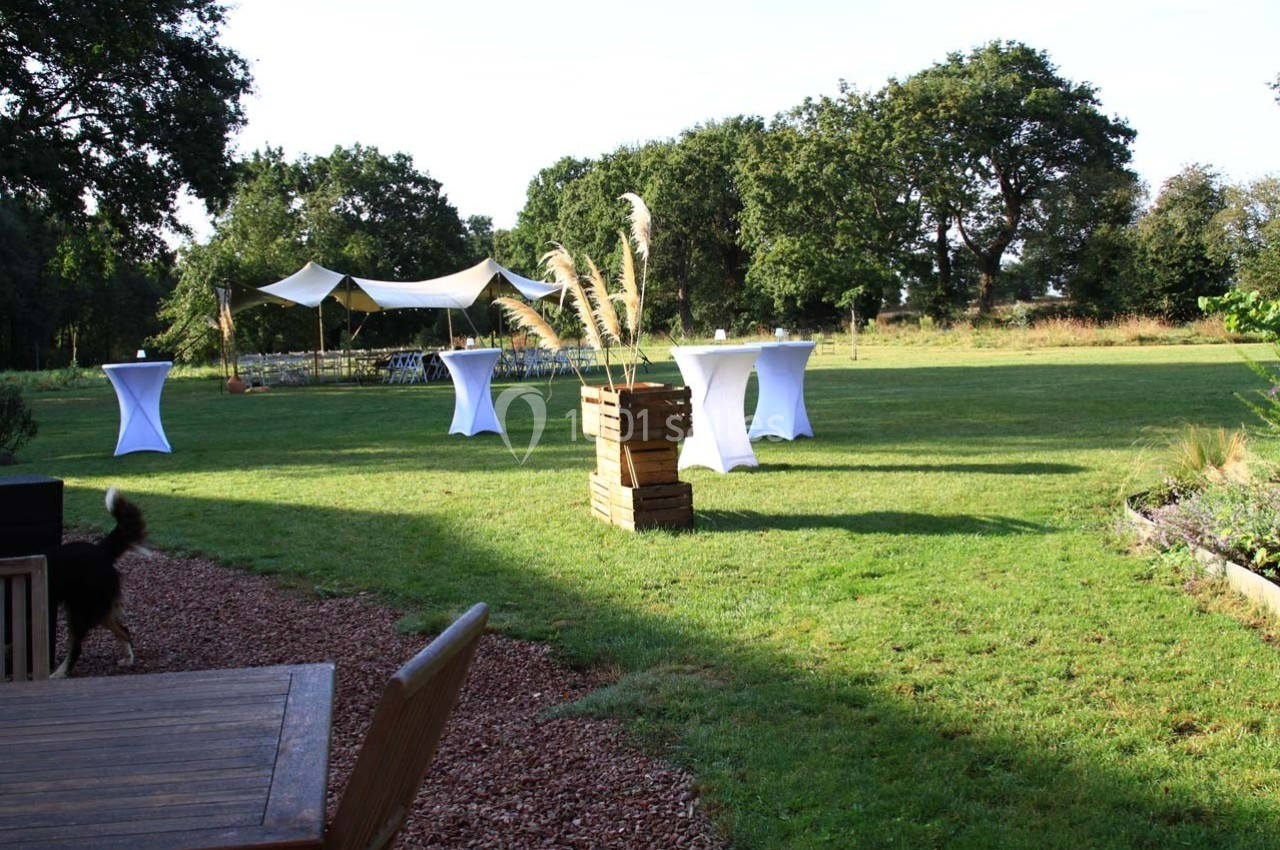 Jardin avec tables hautes et tente blanche en arrière-plan, aménagé pour un événement en plein air.