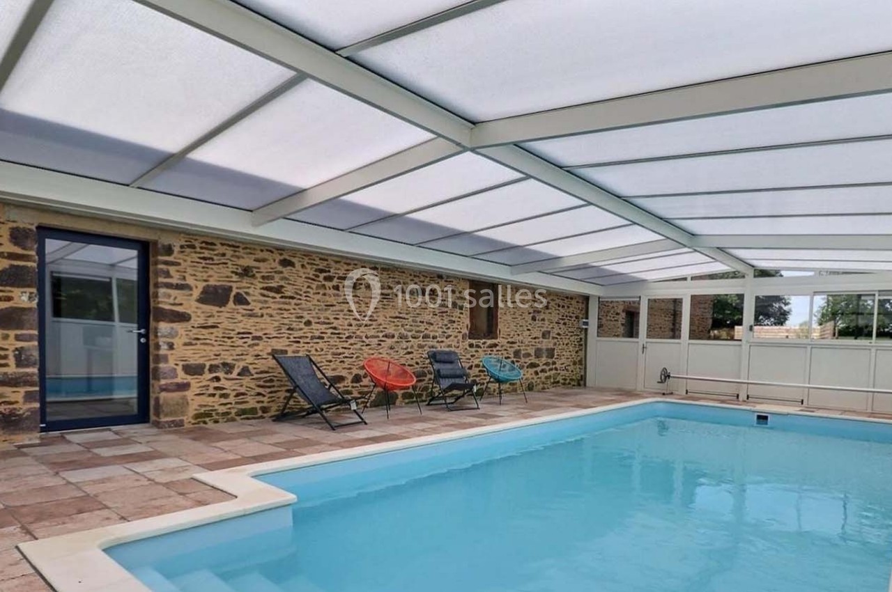 Piscine intérieure avec toit transparent, entourée de murs en pierre et de chaises colorées sur un sol carrelé.