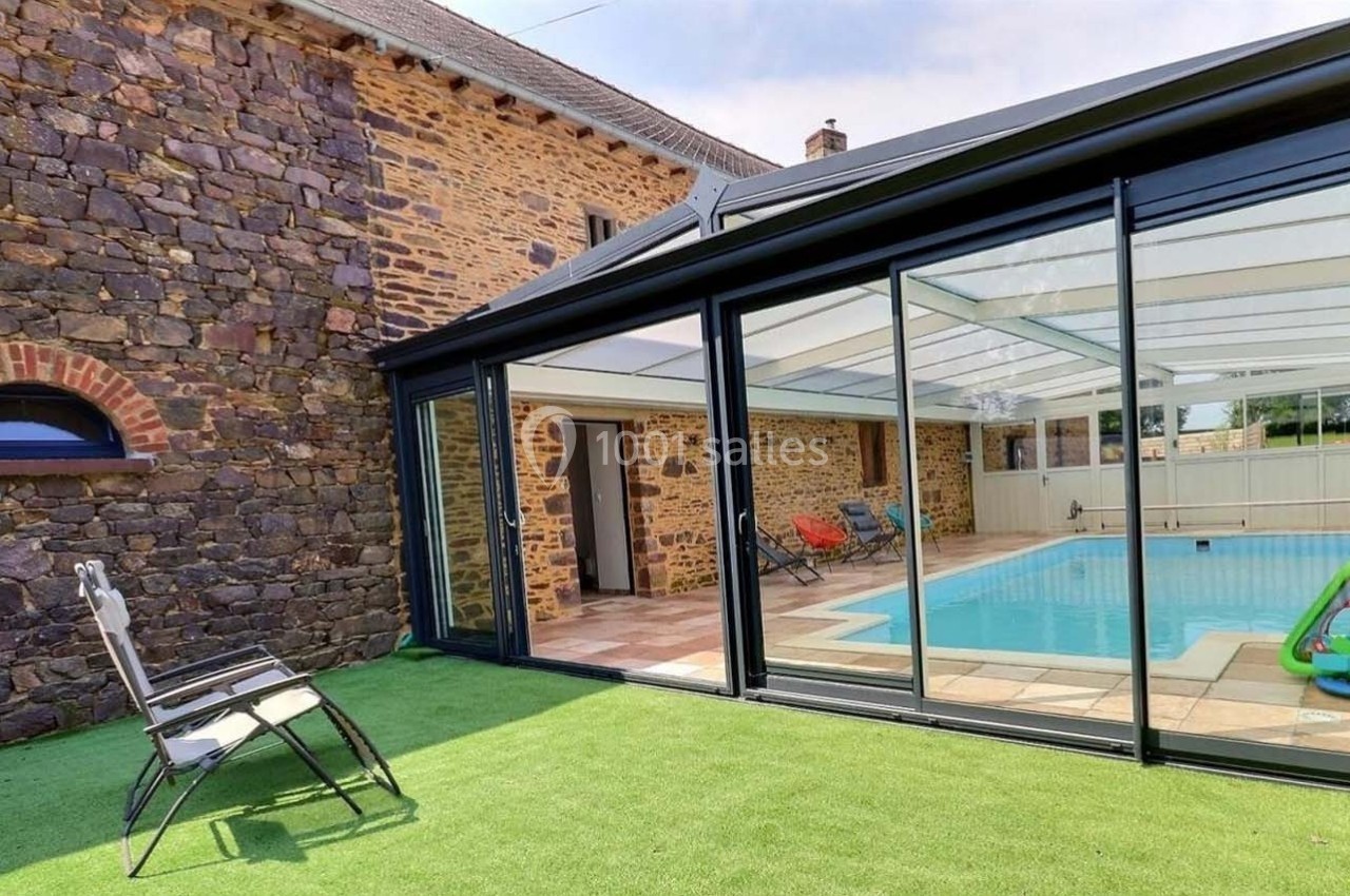 Piscine intérieure avec verrière, entourée d'un mur en pierre et d'une pelouse synthétique, chaise au premier plan.