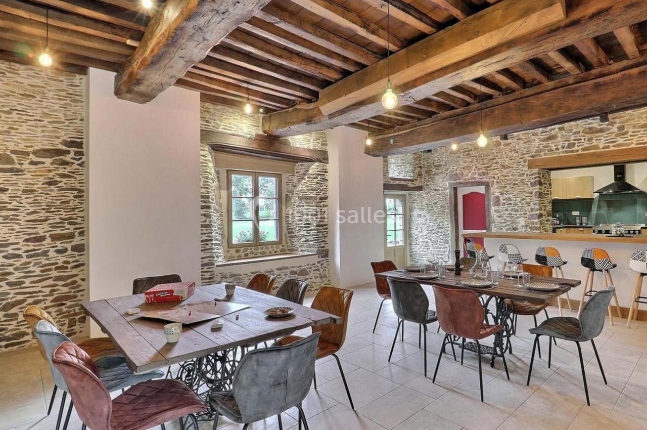 Salle à manger spacieuse avec murs en pierre apparente, poutres en bois, grandes tables et chaises colorées.