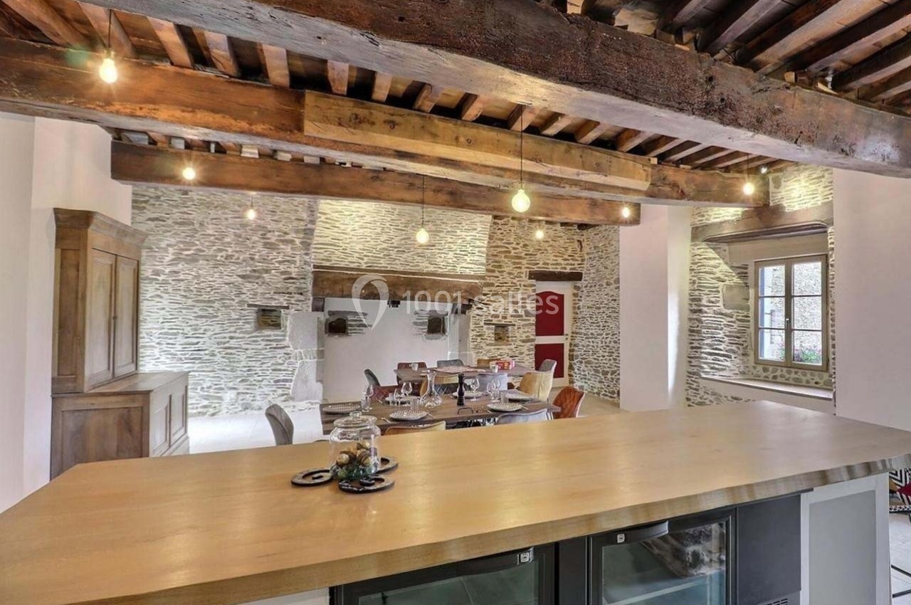 Cuisine ouverte avec un comptoir en bois, poutres apparentes, murs en pierre et table à manger dans un décor rustique.