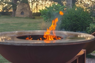 Brasero en métal allumé dans un jardin, entouré de végétation et d'arbres au coucher du soleil.