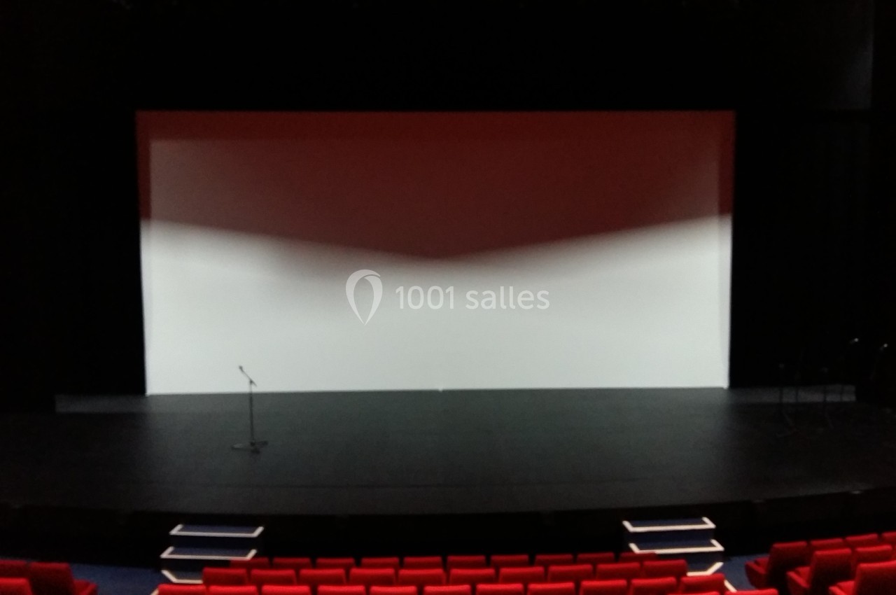Scène de théâtre vide avec un écran blanc en fond, un micro sur pied et des sièges rouges au premier plan.