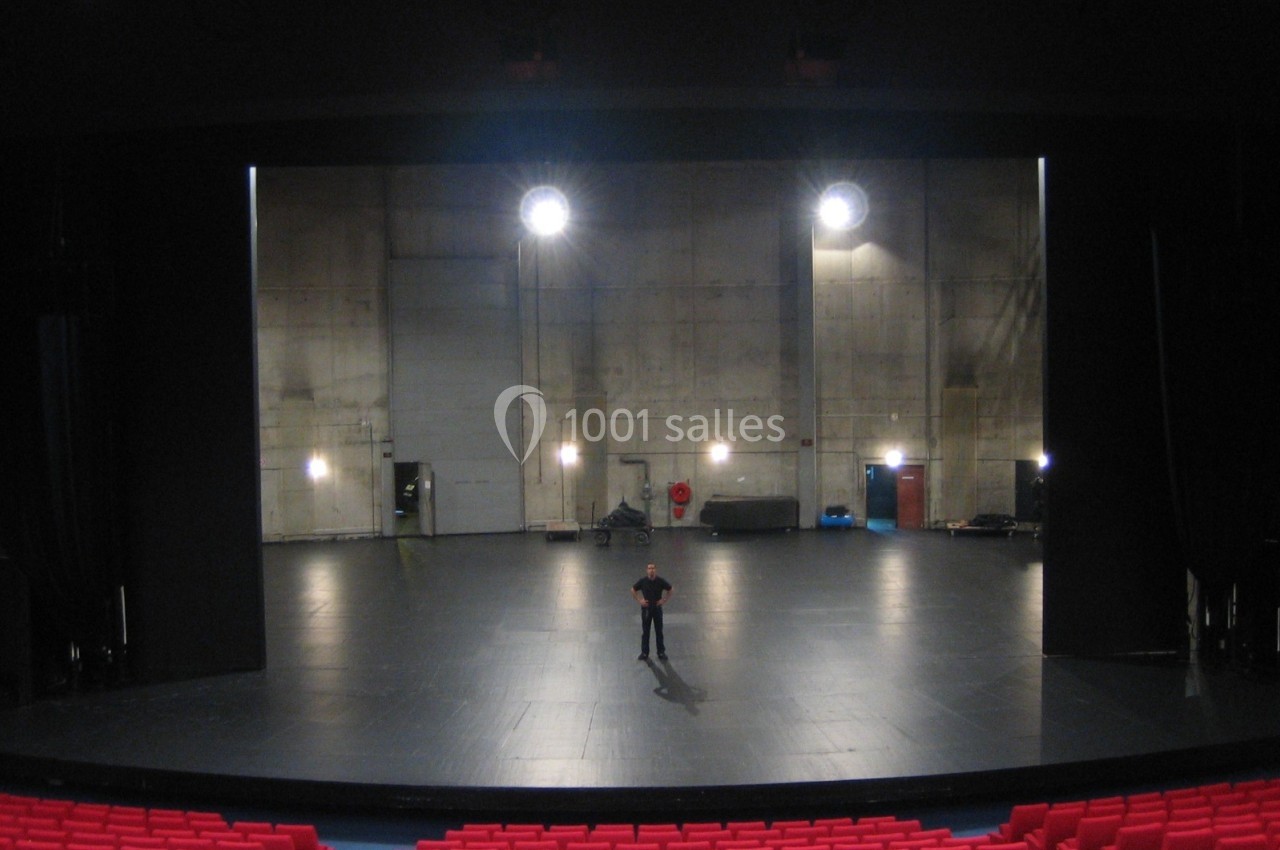 Une scène de théâtre vide vue depuis la salle, avec un homme debout au centre et des sièges rouges au premier plan.