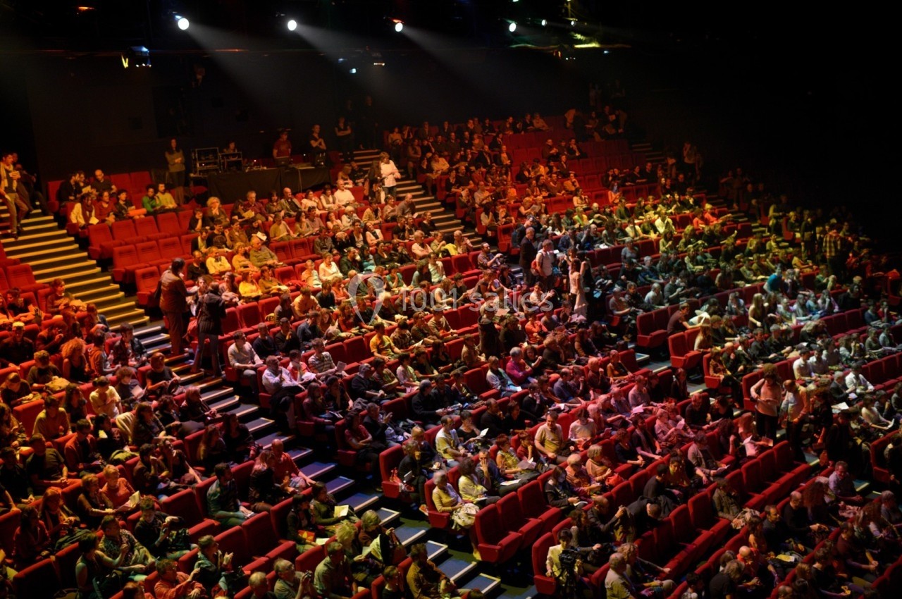 Salle de spectacle remplie de spectateurs assis, éclairée par des projecteurs colorés.