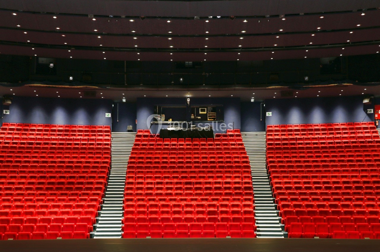 Salle de théâtre avec des rangées de sièges rouges vides et une scène vue depuis le public.