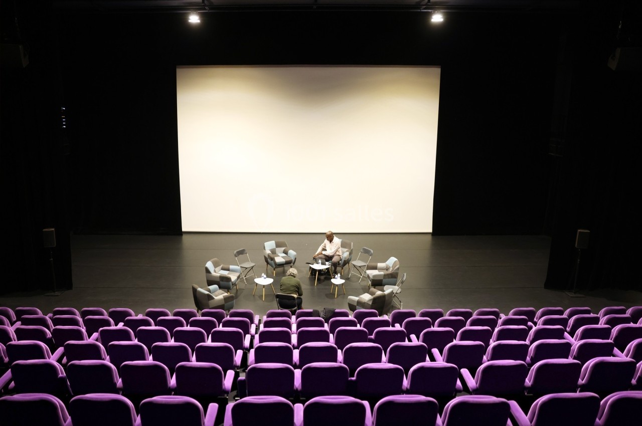 Salle de conférence vide avec des sièges violets, un écran blanc et quelques fauteuils disposés sur la scène.