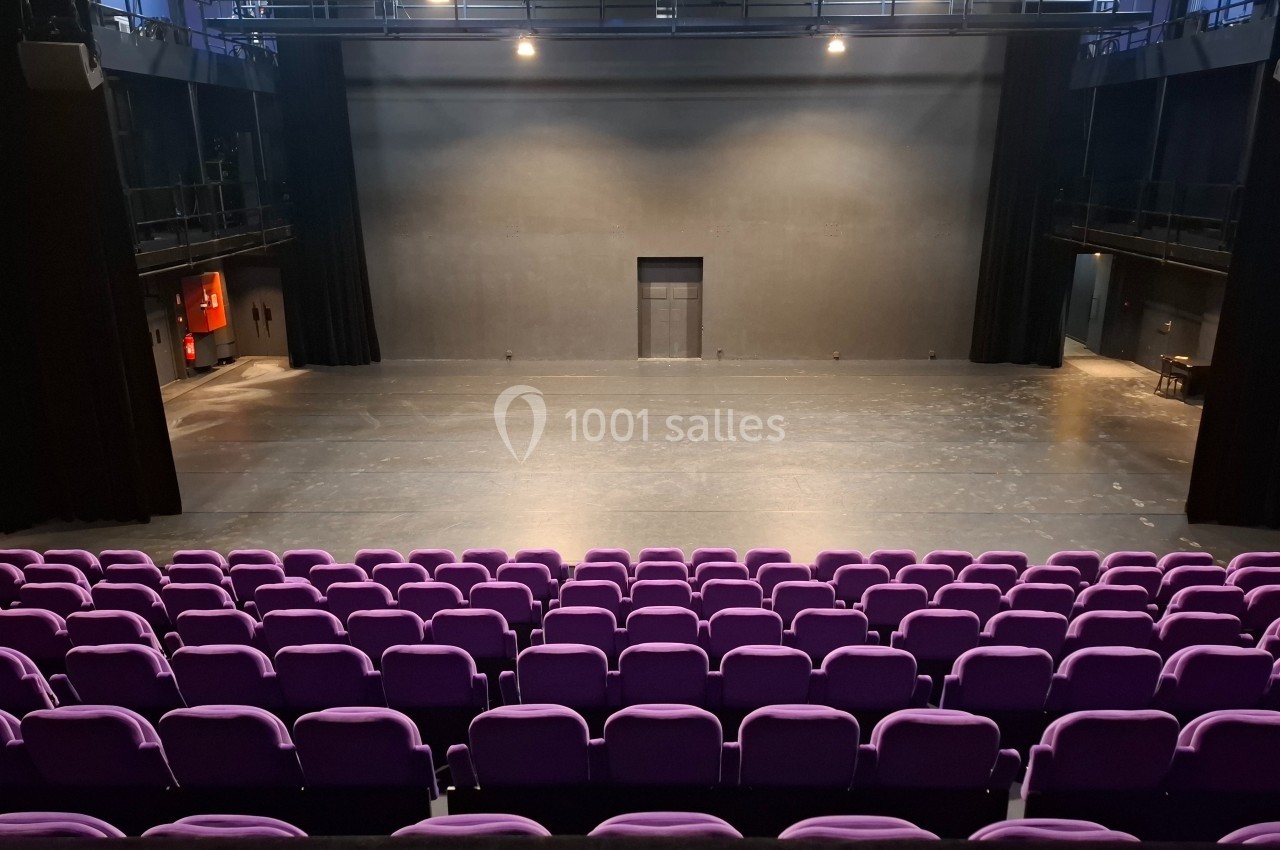 Salle de théâtre vide avec des rangées de sièges violets et une scène éclairée par des projecteurs.