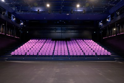 Salle de spectacle vide avec des rangées de sièges violets face à une scène sombre.