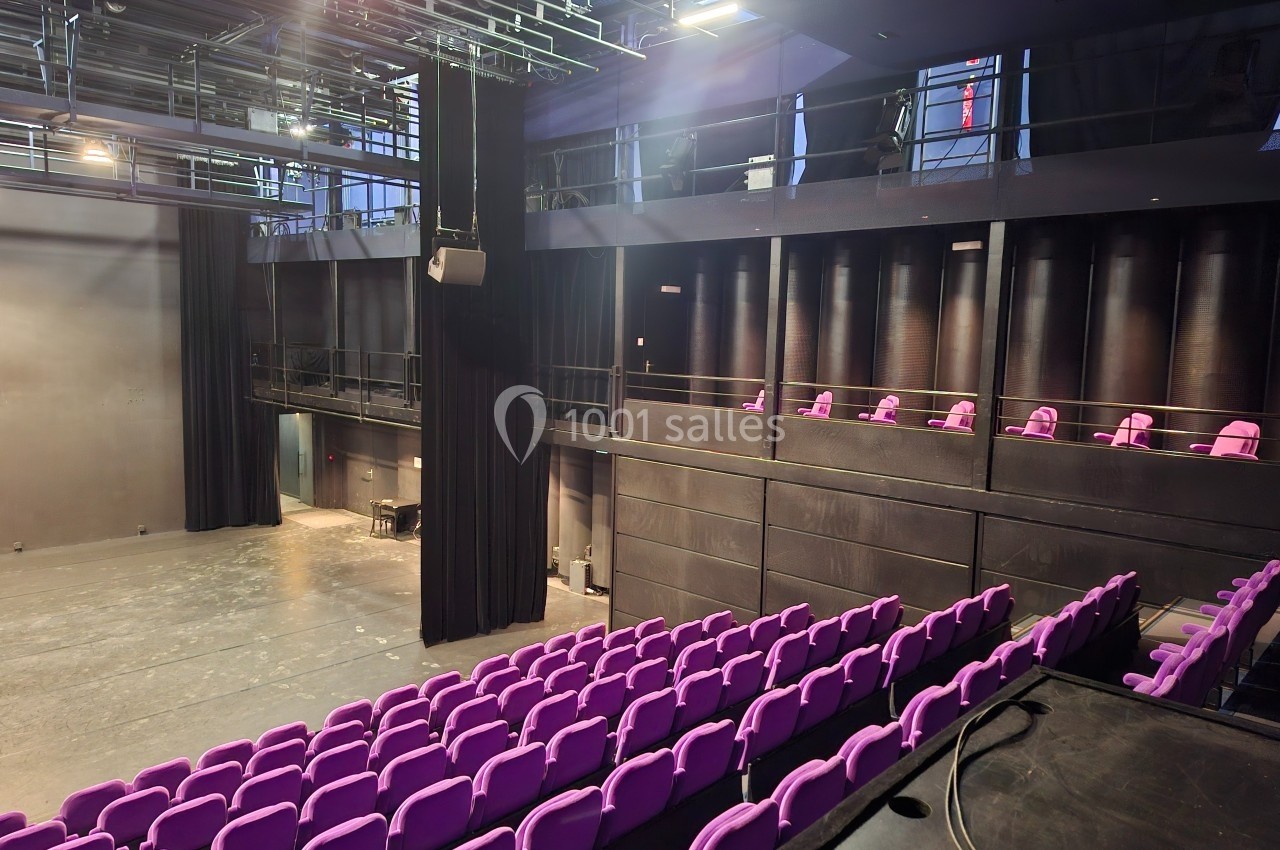 Salle de spectacle avec sièges violets, scène vide et balcons latéraux, éclairée par des projecteurs.