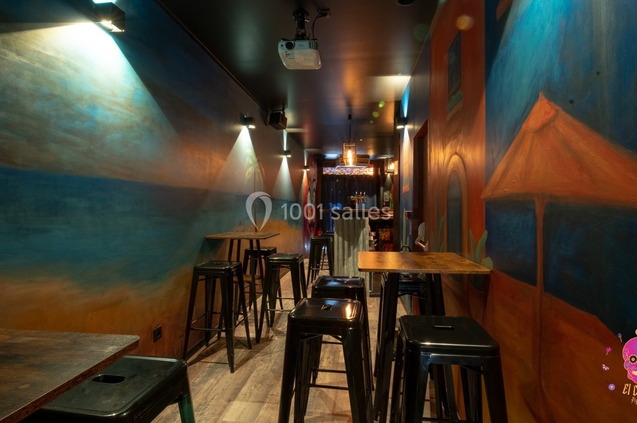Salle étroite avec tables hautes et tabourets, décorée de fresques colorées et éclairée par des lumières tamisées.