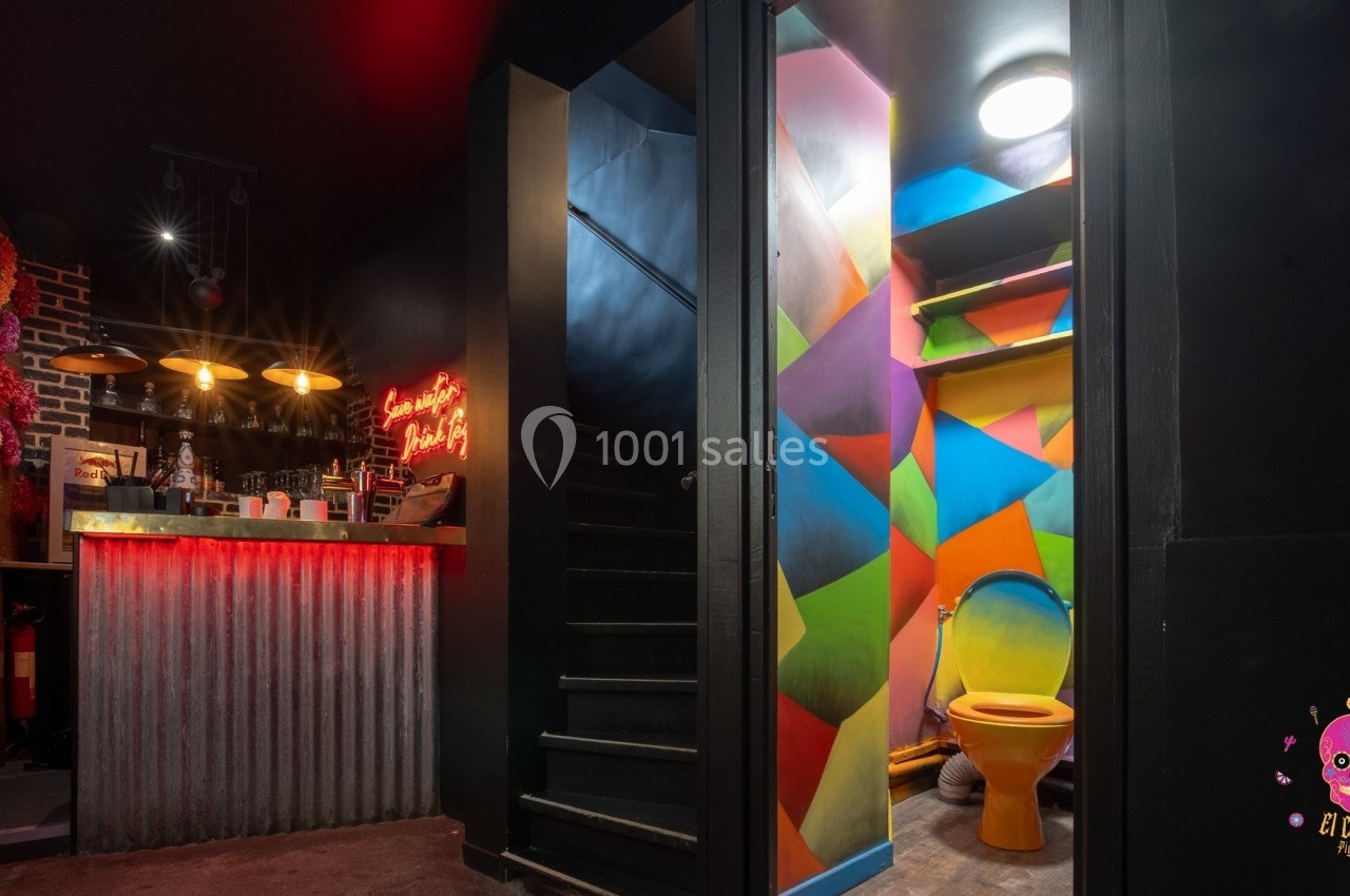 Vue d'un bar avec éclairage tamisé, un escalier sombre et des toilettes aux murs colorés et vifs.