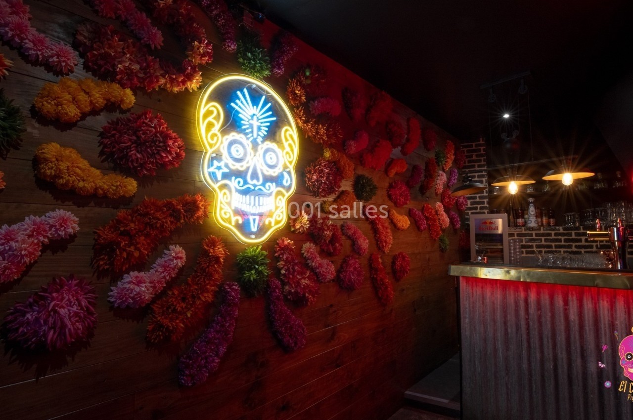 Mur décoré de fleurs colorées et d'une enseigne néon en forme de crâne, avec un bar éclairé en arrière-plan.