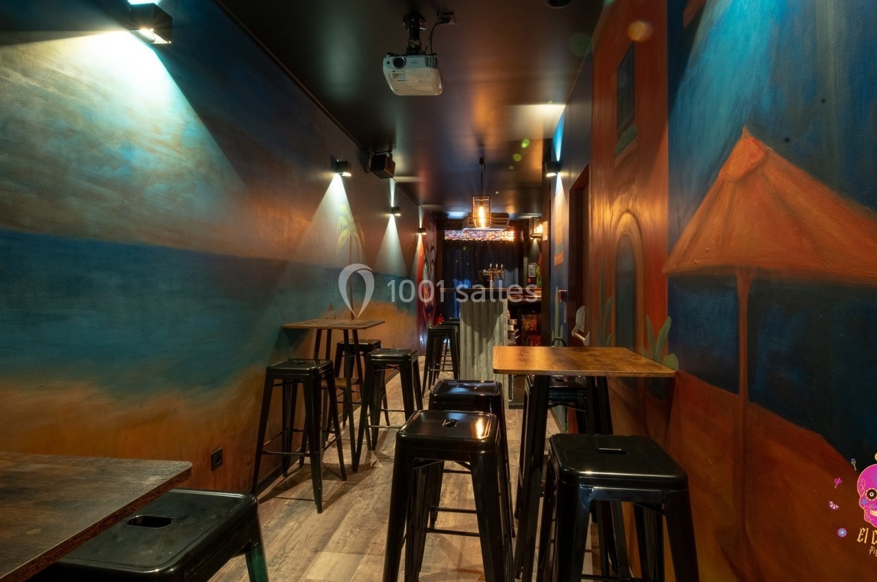 Salle étroite avec tables hautes, tabourets noirs, murs peints colorés et éclairage tamisé.