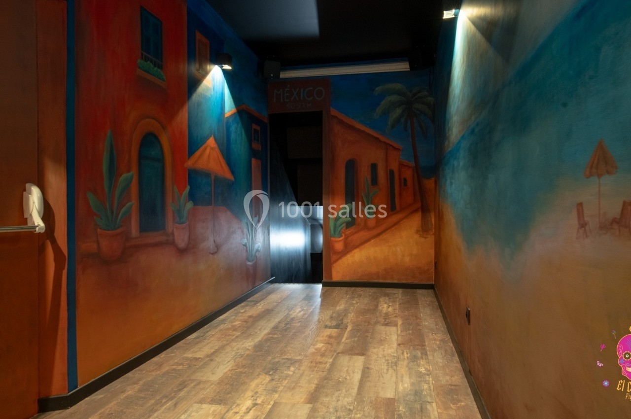 Murales colorées représentant une rue et une plage mexicaines dans une pièce au sol en bois, éclairée par des spots.