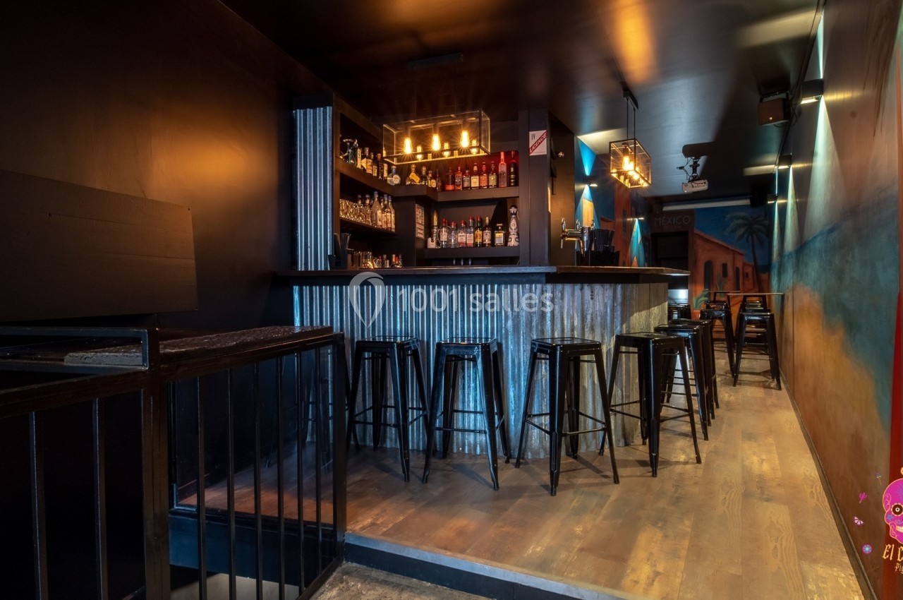 Bar à l'ambiance tamisée avec comptoir en métal, tabourets hauts et étagères garnies de bouteilles.