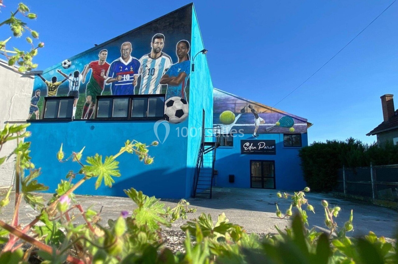 Fresque murale colorée représentant des sportifs sur la façade d'un bâtiment bleu, avec des plantes au premier plan.