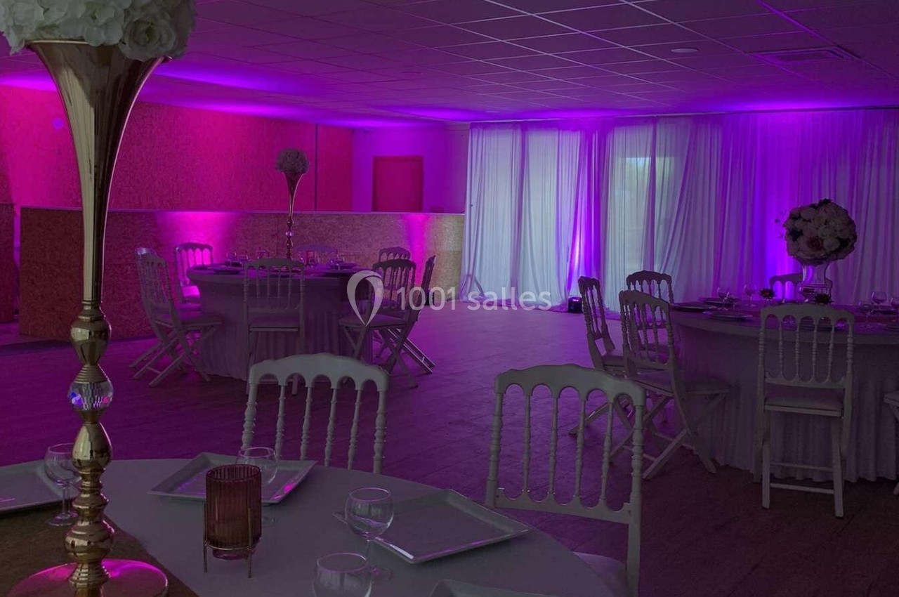 Salle de réception décorée avec des tables rondes, nappes blanches et éclairage d'ambiance rose-violet.