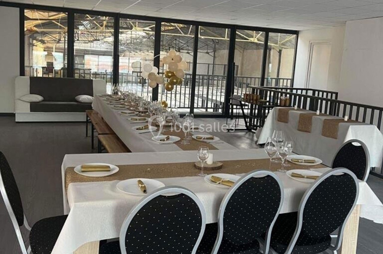 Salle de réception aménagée avec une longue table dressée, nappes blanches, décorations dorées et chaises noires.