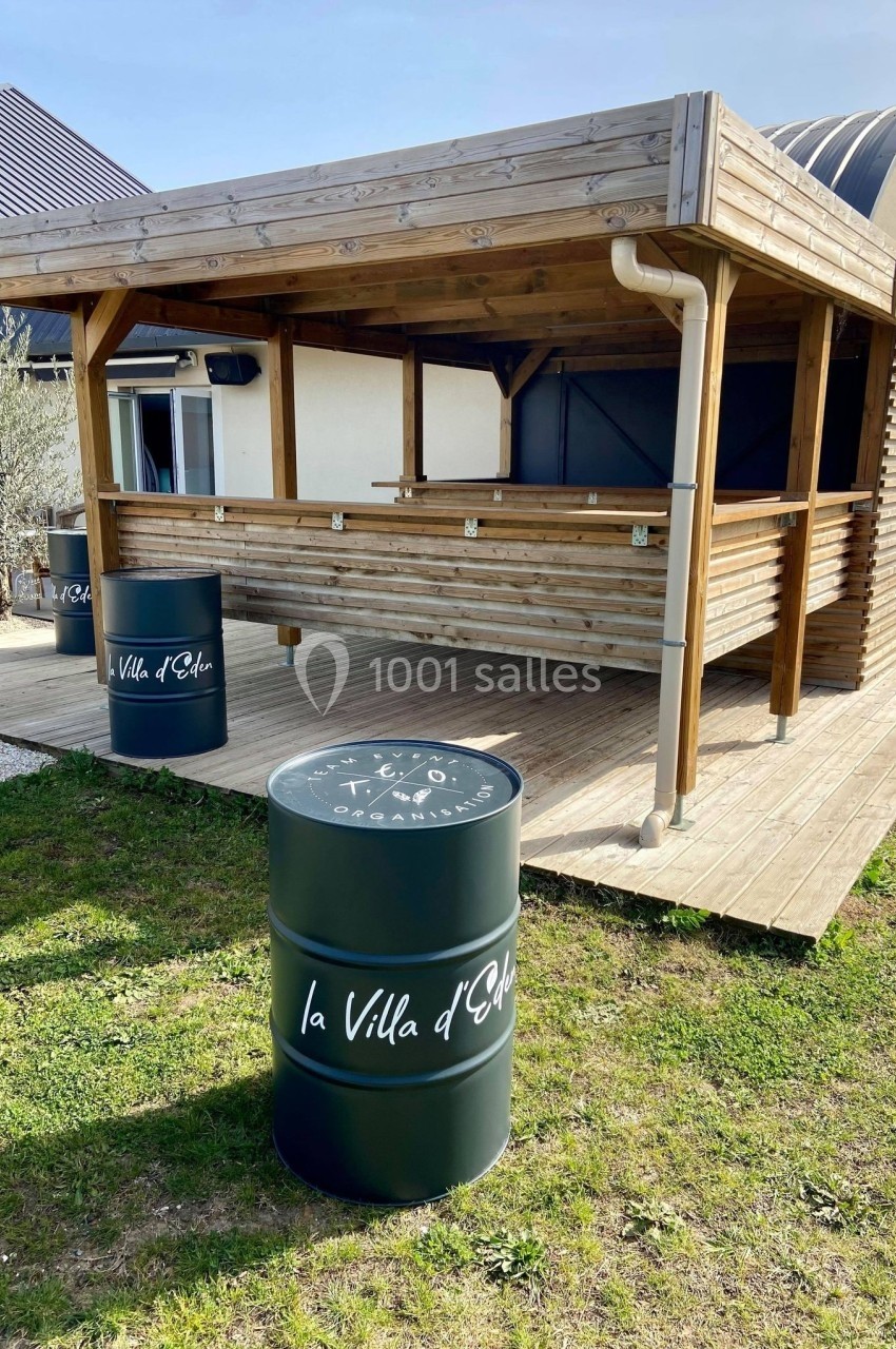 Terrasse en bois avec un comptoir couvert et deux tonneaux noirs marqués ’la Villa d'Elba’ sur une pelouse.