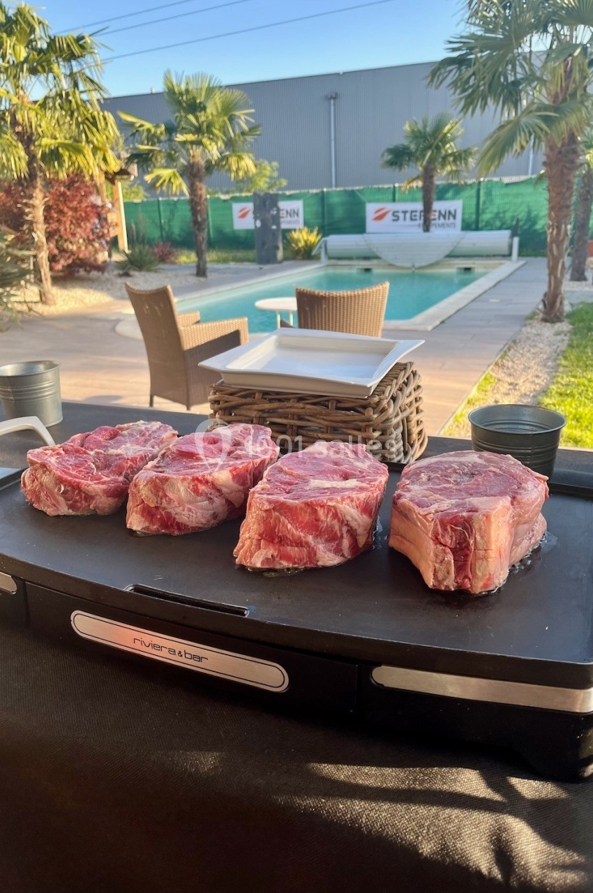 Quatre morceaux de viande rouge crue posés sur une plancha, en extérieur près d'une piscine et de palmiers.