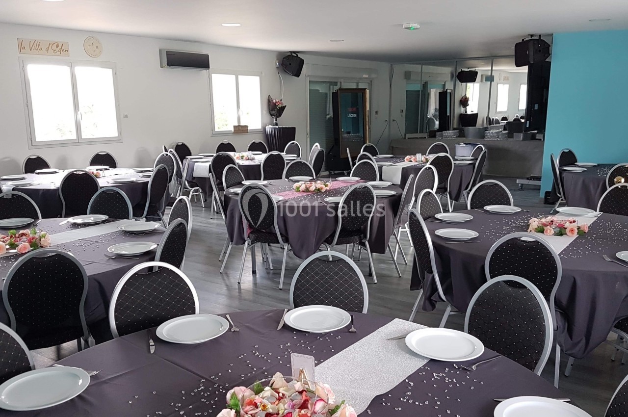 Salle de réception décorée avec des tables rondes, nappes grises, assiettes blanches et arrangements floraux.